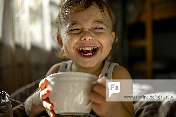 Cute baby with cup laughing in bed at home
