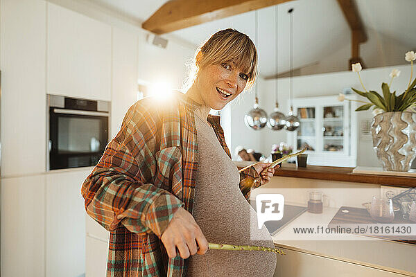 Happy expectant woman playing with asparagus sticks in kitchen at home