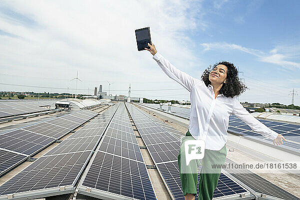 Smiling businesswoman with arms outstretched holding tablet in front of solar panels