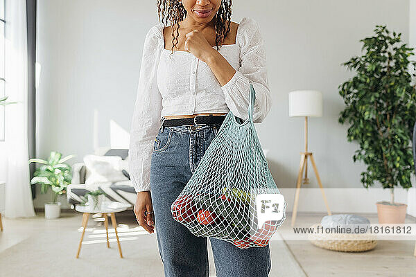 Young woman carrying mesh bag with vegetables at home