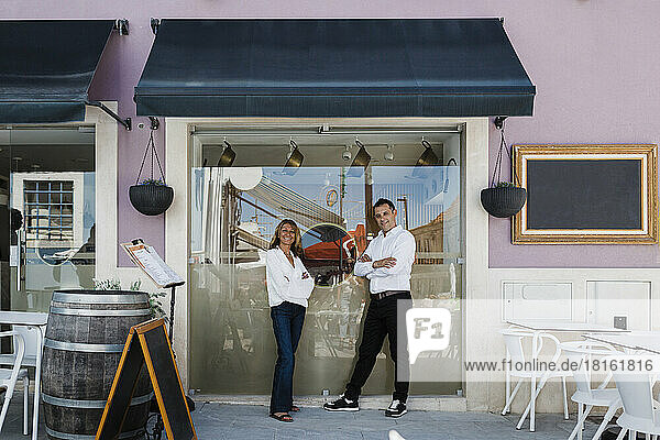 Smiling restaurant owners with arms crossed standing by glass window