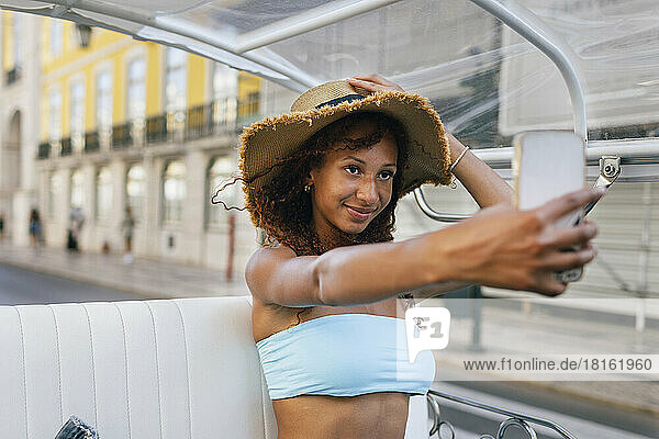 Smiling woman wearing hat taking selfie through smart phone sitting in tuk tuk