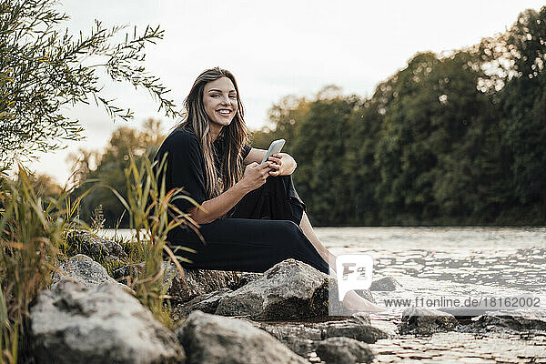 Smiling woman with smart phone sitting by lake