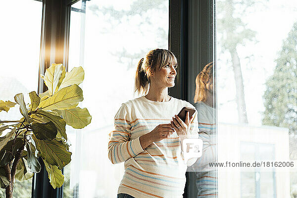 Smiling pregnant woman holding mobile phone leaning on glass door