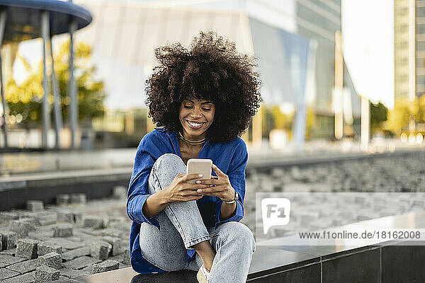 Smiling Afro woman using smart phone sitting on wall