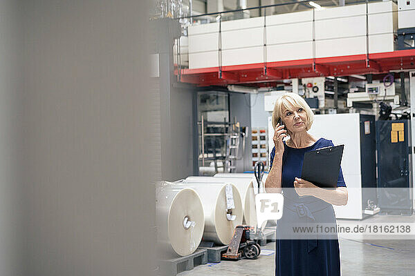 Senior businesswoman holding clipboard talking on mobile phone in factory
