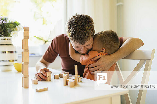 Son embracing father by stacked toy blocks on table at home