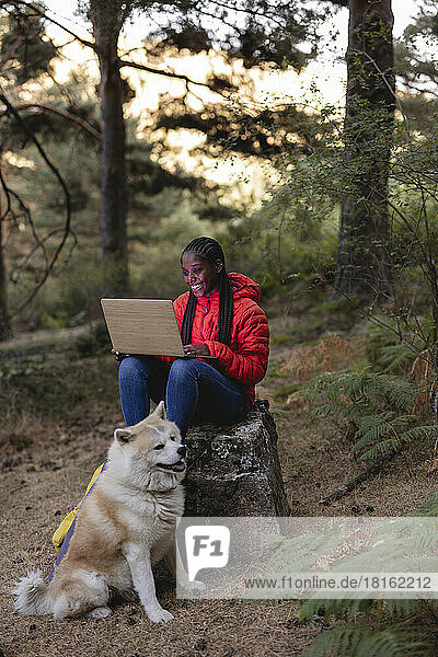 Happy woman using laptop with dog sitting in forest