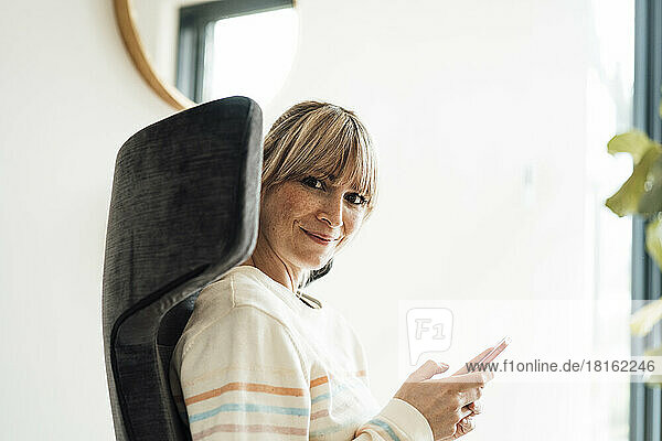 Lächelnde Frau sitzt mit Smartphone auf einem Stuhl
