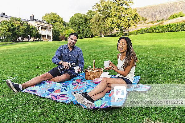 Multiracial couple enjoying drinks sitting on picnic blanket in lawn
