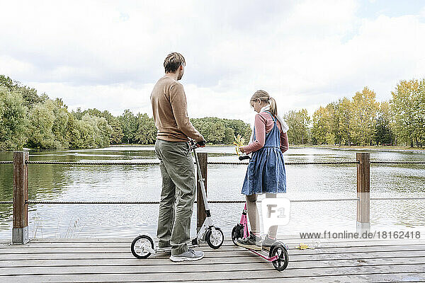 Vater und Tochter mit Tretrollern auf Fußgängerbrücke über See im Park