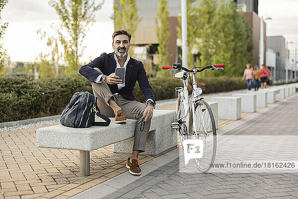 Smiling businessman with smart phone sitting on bench by bicycle