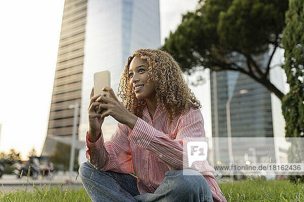 Smiling woman with smart phone contemplating on grass in city