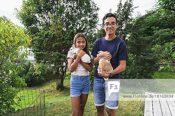 Brother and sister holding rabbits standing in back yard