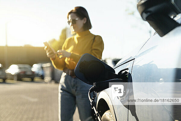 Electric car charging at station with woman using smart phone in background on sunny day