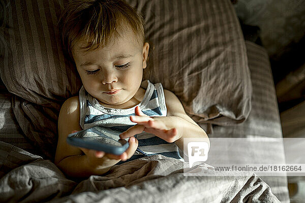 Cute toddler using smart phone lying in bed in bedroom