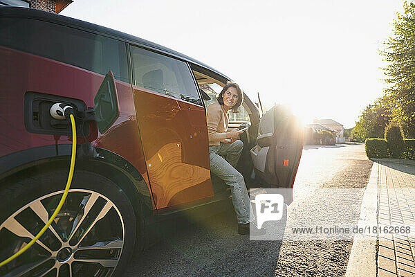 Glückliche Frau mit Mobiltelefon sitzt an einem sonnigen Tag im Elektroauto an der Fahrzeugladestation