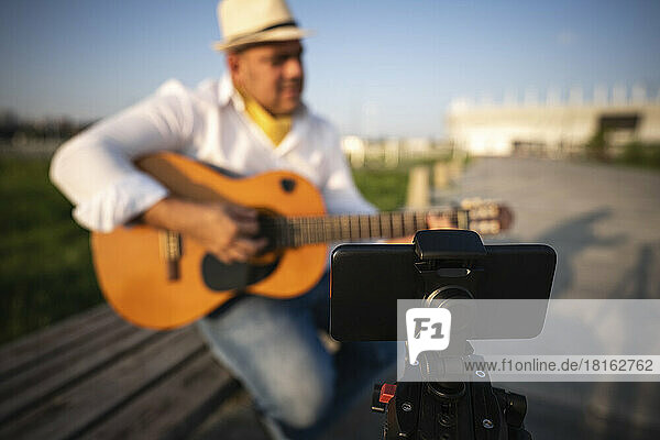 Street musician playing guitar and recording through smart phone on tripod