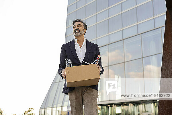 Lächelnder Geschäftsmann verlässt das Büro mit Büromaterial im Karton
