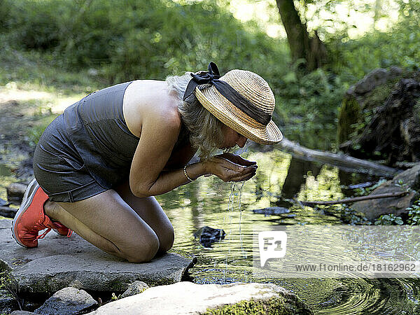 Thirsty senior woman wearing hat drinking water from stream in forest