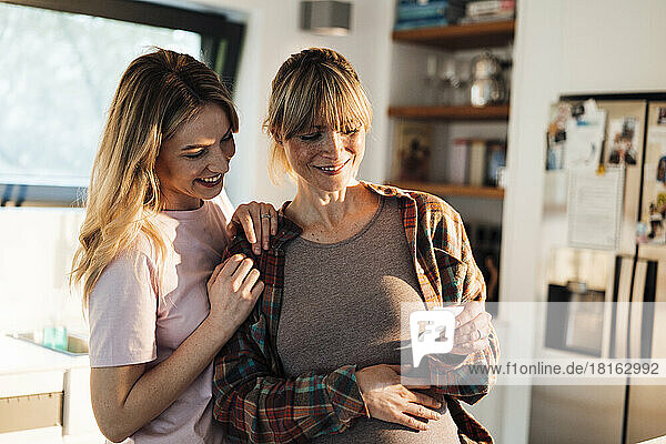 Smiling expectant woman showing ultrasound to sister at home