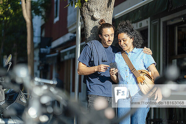 Woman sharing mobile phone with boyfriend by tree trunk