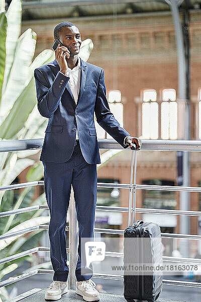 Smiling businessman with luggage talking on smart phone by railing at railroad station