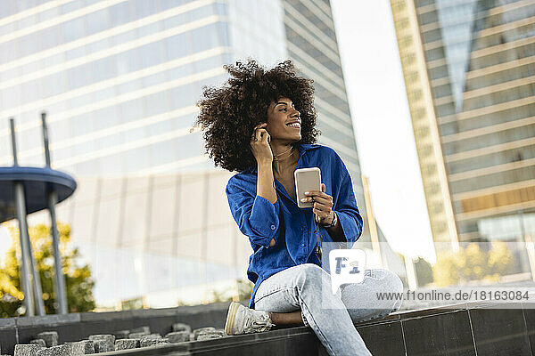 Happy Afro woman holding mobile phone sitting on wall
