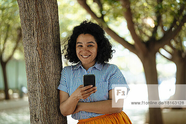 Happy woman with mobile phone leaning on tree trunk