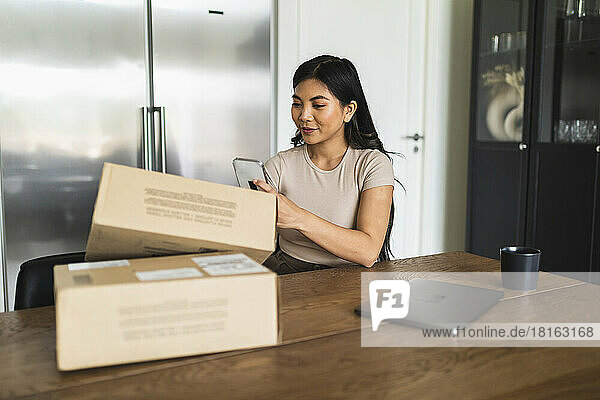 Young woman taking picture of delivery package box at dining table