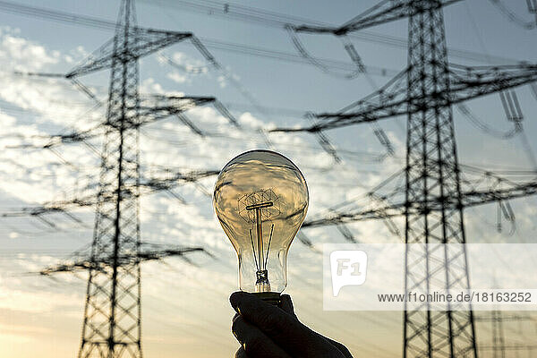 Woman's hand holding light bulb in front of electricity pylons