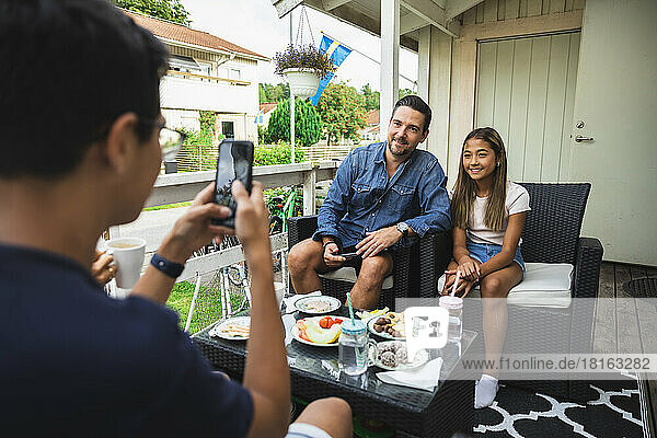 Boy photographing father and sister through smart phone sitting on chair at porch
