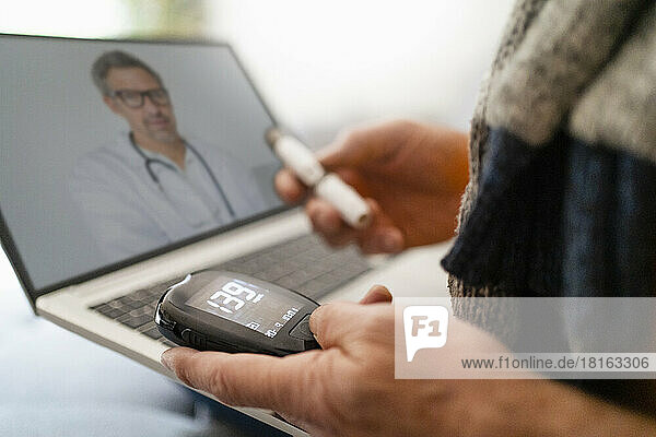 Man measuring blood sugar through device doing online consultation with doctor on laptop
