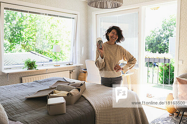 Happy woman holding rolled up clothes standing with hand on hip in bedroom