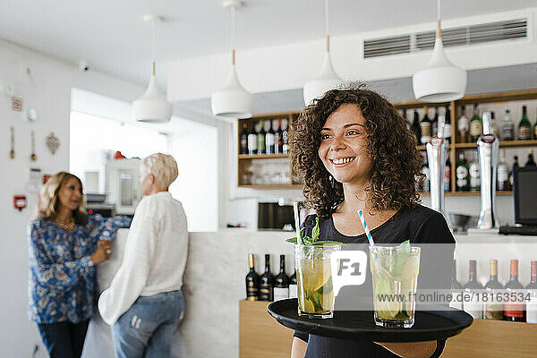 Happy young waitress with tray serving cocktail drinks at restaurant