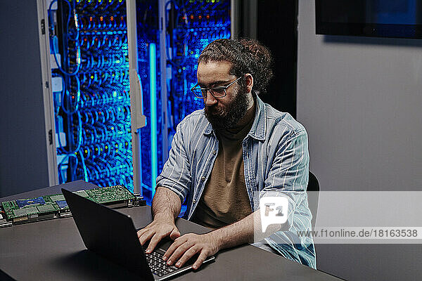 IT expert working on laptop sitting at desk in server room