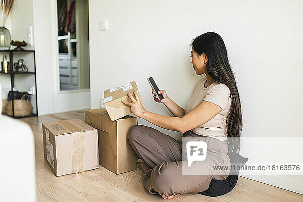 Young woman checking and taking picture of delivery package box through smart phone at home