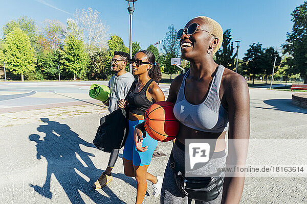 Smiling sportswoman with basketball walking by friends on footpath
