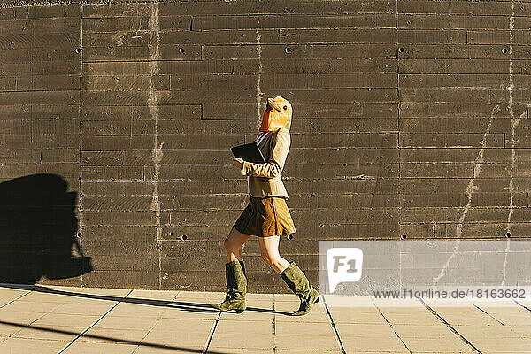 Freelancer wearing bird mask walking with tablet PC by wall