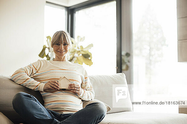 Lächelnde schwangere Frau mit Hausmodell sitzt zu Hause auf dem Sofa