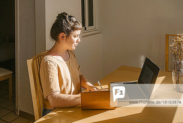 Graphic designer writing in book with laptop at desk