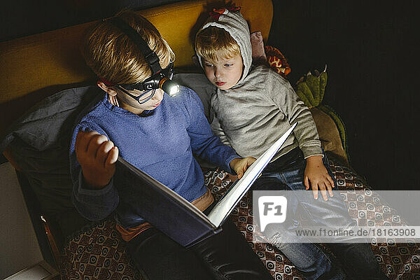Boy wearing headlight reading book with brother on bed at home