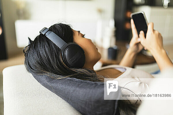 Woman wearing wireless headphones and using smart phone on sofa