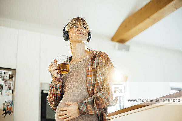 Smiling pregnant woman with headphones holding tea mug at home