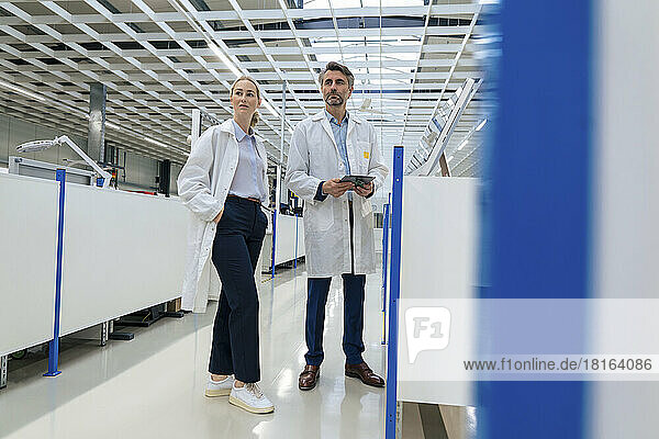 Engineer with hands in pockets standing by colleague holding tablet PC at factory