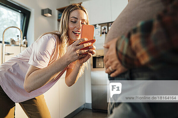 Woman photographing pregnant belly of sister through smart phone at home