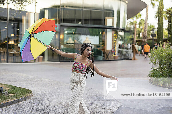 Young woman with arms outstretched holding colorful umbrella on footpath