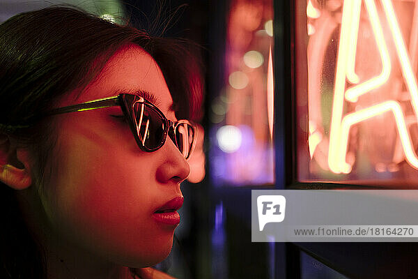 Young woman wearing sunglasses by illuminated wall with neon lights