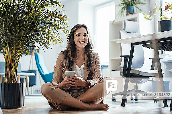 Happy young businesswoman with mobile phone sitting in home office