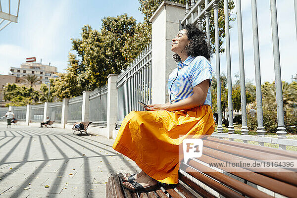 Lächelnde Frau genießt das Sonnenlicht  während sie auf einer Bank im Park sitzt